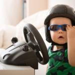 bigstock–140228711 Cute little kid boy in a helmet playing with Computer steering wheel. Future driver. Getting ready for professional driving. Child driving a car. Funny toddler boy indoors. Boy and a car.